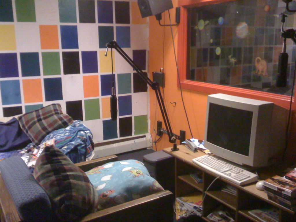 Haverford College radio station WHRC's lounge/live music space with view of Studio B in 2009. Wall with brightly painted squares in the background and an orange-framed window that has planet and dinosaur stickers on it to the right. A table holds a desktop computer and there's a microphone attached to the table with a boom arm. A wood-framed chair has plaid and Sponge Bob pillows and there appears to be a couch behind that. Photo: J. Waits