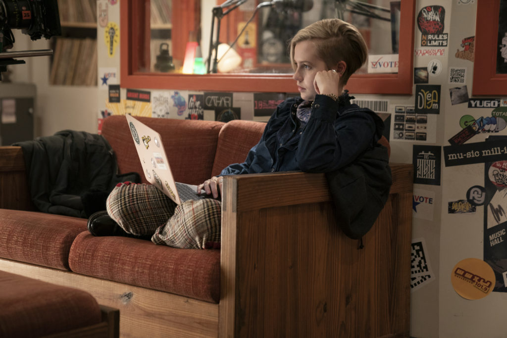 Angourie Rice (as Siobhan Sheehan) at fictional Haverford College radio WWXU on Mare of Easttown. She sits on an orange, wood-framed couch holding a sticker-covered laptop. Behind her we see the window frame of the DJ booth. The walls are covered with stickers, including one for WWXU 101.9 FM. Photograph by Sarah Shatz/HBO