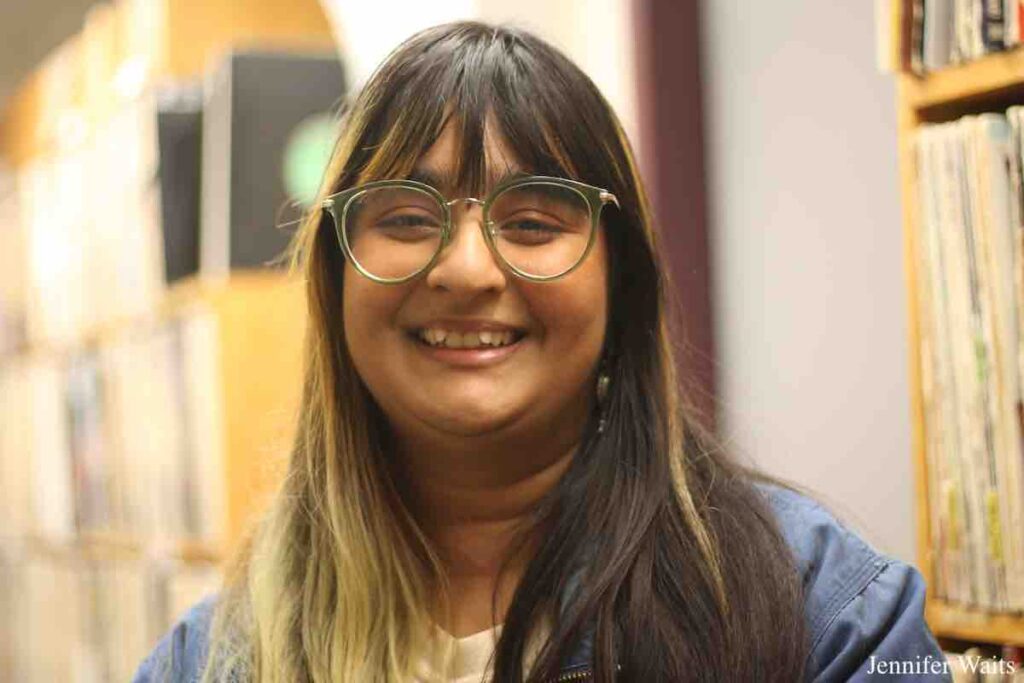 College radio station WCDB's General Manager Mehr Sharma standing in front of several shelves of vinyl records. Image of person with long brown and blonde hair, wearing glasses. Photo: J. Waits