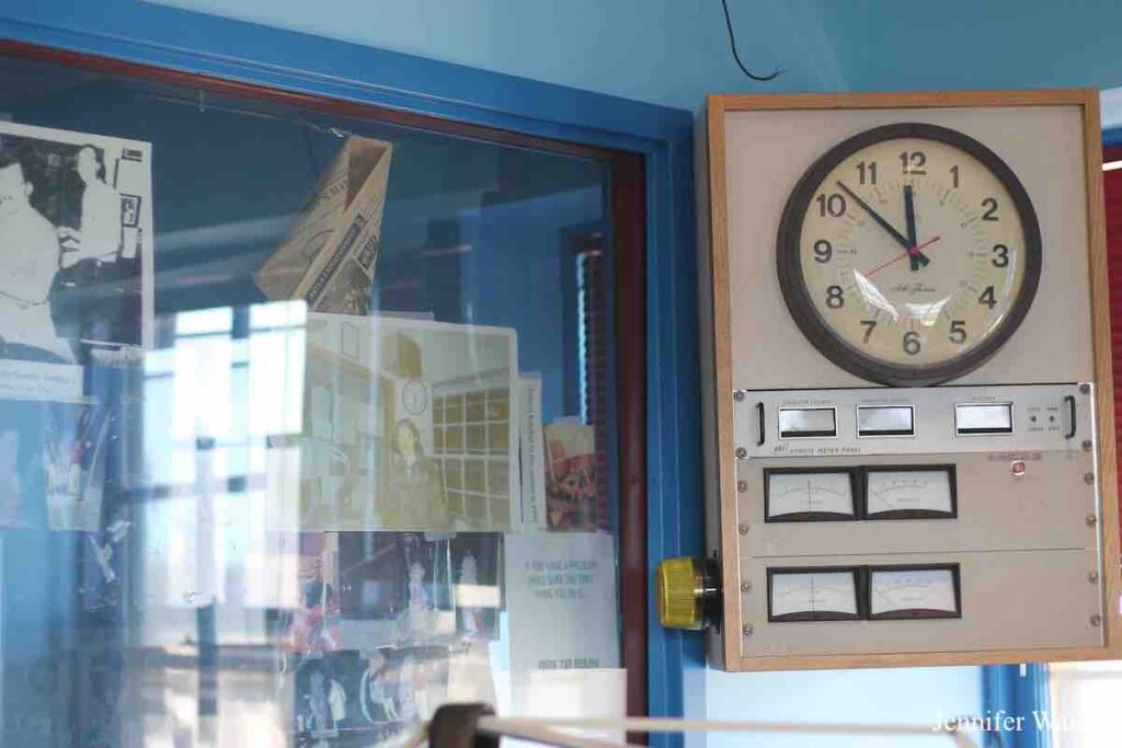 Photo of college radio station WRUC's upstairs studio. A large clock is on the upper right, with various meters below it. To the left is a window looking into another studio. Old photos are taped to the window, along with some flyers. Walls of room are baby blue, with window framed in a deeper blue color. Photo: J. Waits