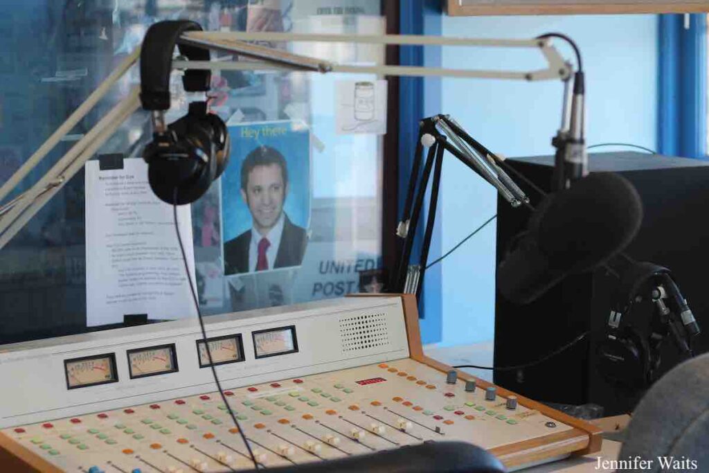 Photo of studio for college radio station WRUC. Mixing board at center, with microphone over it, hanging on a boom. Black headphones are hanging on the boom. A window looks into another studio and there are flyers and a photo of a man posted on that window. Photo: J. Waits