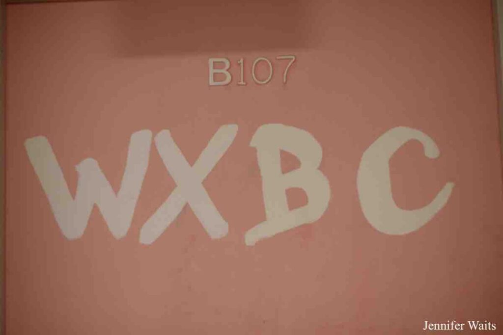 Photo of door to college radio station WXBC. Pink background with the number B107 above handwritten letters WXBC in white. Photo: J. Waits