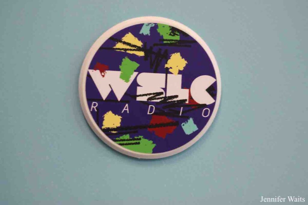 Photo of WSLC sticker on a light blue wall. Sticker has purple, black, yellow, blue, green, and red abstract designs in front of the letters WSLC in white. Photo: J. Waits