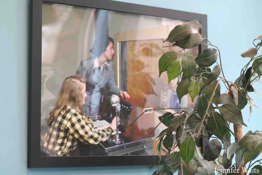 Photo at college radio station WSLC. Light blue walls with black framed photo from late 40s or early 1950s of three women in a radio studio doing radio. Plant in front of photo. 2023 photo by J. Waits