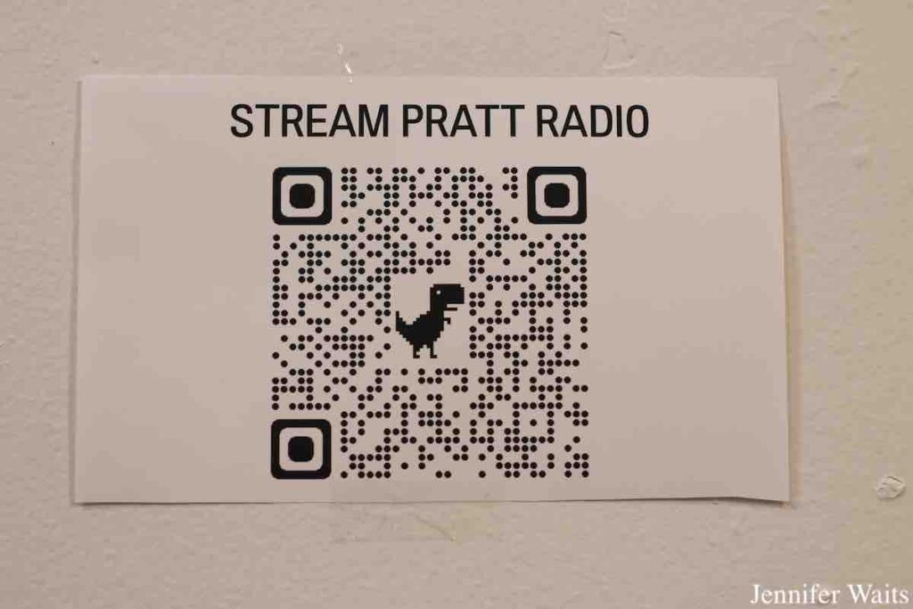 Flyer on wall at WPIR Pratt Radio in March, 2023. Flyer reads "Stream Pratt Radio" and has large QR code  on it with a dinosaur in the middle. Photo: J. Waits