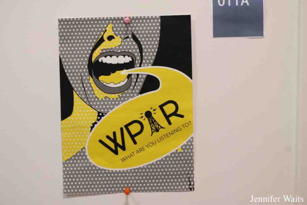 WPIR Poster on wall at college radio station WPIR Pratt Radio in March, 2023. Poster is black, grey, yellow and white, with "WPIR What are You Listening To" in a speech bubble coming out of a drawing of an open mouth. Photo: J. Waits