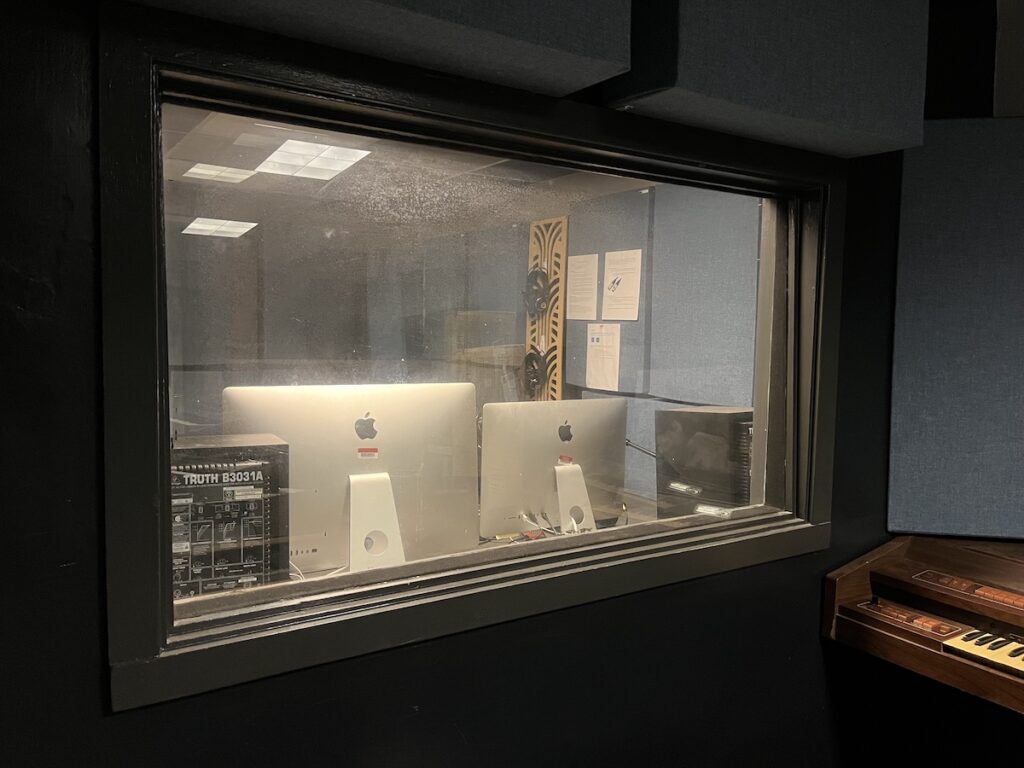 Photo of Haverford College radio station WHRC in spring 2023. In the photo, there's a large window looking into the radio room. The backs of two Apple monitors can be seen, as well as headphones hanging on a wall. Photo: Natalie White