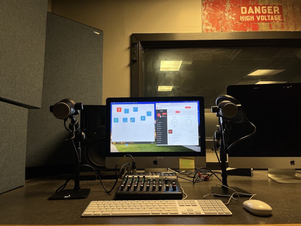 Haverford College radio station WHRC in spring, 2023. In photo: a computer monitor with microphones in front of it. There's also a mixing board and a computer keyboard with a mouse. In the background is a large window. Above it is a sign that reads "danger high voltage." Photo: Natalie White