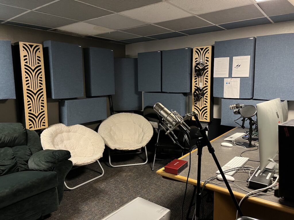 Photo of Haverford College radio station WHRC in spring, 2023. Shown: a microphone on a stand, a desk with computers, keyboards, microphones, and a pop screen. The room also has a couch and two round chairs. Headphones hand on a wall. Photo: Natalie White