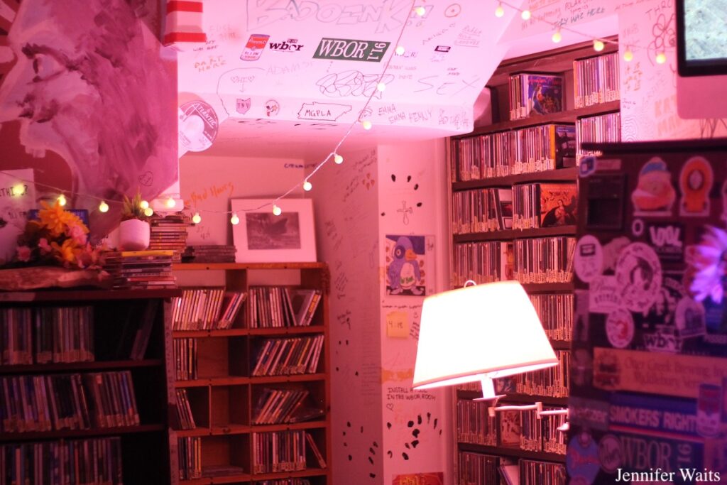 WBOR at Bowdoin College. Pictured: shelves of CDs and walls covered with posters, writing, drawings and stickers. Sticker-covered furniture is in the front of the photo, next to a swing-arm lamp. Photo: J. Waits