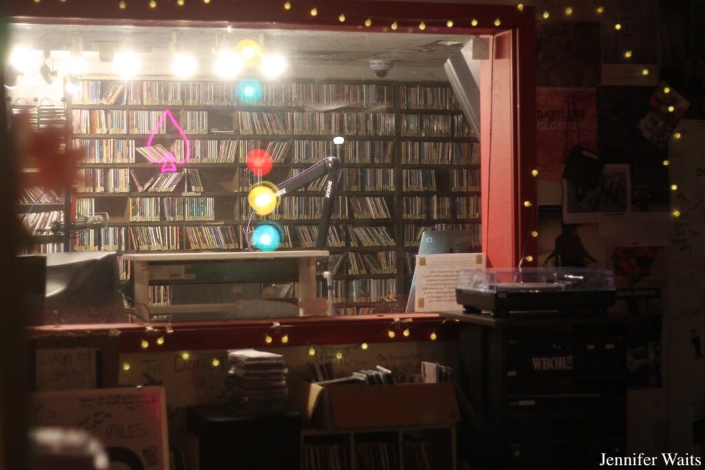 Photo of Bowdoin College radio station WBOR. Records on shelves in the distance, turntable to the right, and reflections from a traffic light and pink neon sign can be seen in station window. Photo: J. Waits