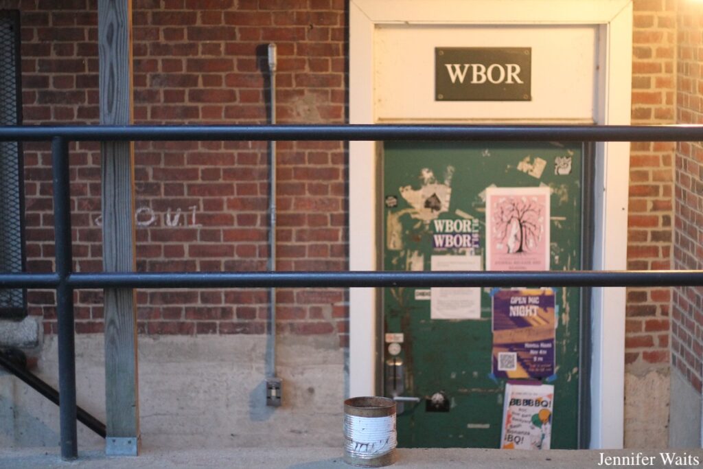 Entrance to Bowdoin College radio station WBOR in August 2024. Pictured: WBOR sign with green door below. Door is covered with posters, WBOR stickers, and other signage. Photo: J. Waits