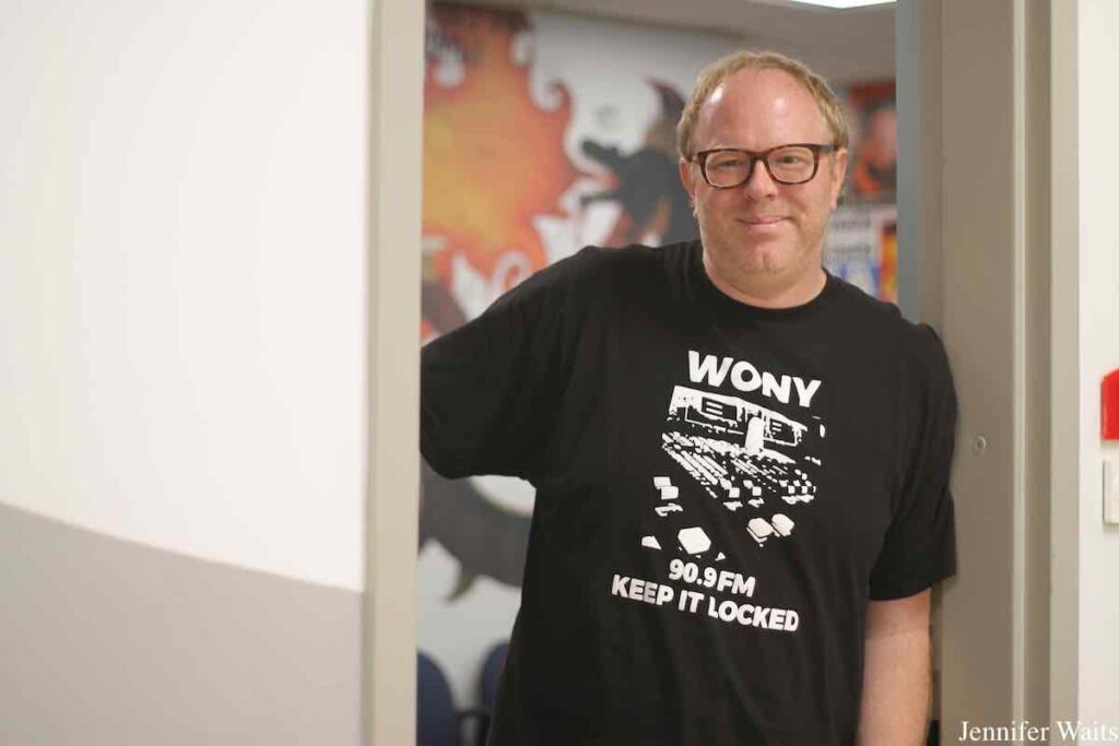 Photo of WONY-FM faculty adviser Andrew Bottomley leaning in a door frame at the college radio station. He's wearing a black Tshirt that reads "WONY 90.9 FM Keep it Locked." Photo: J. Waits