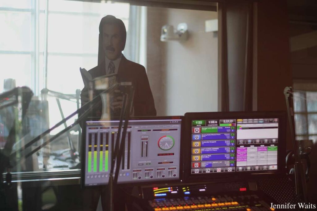 Photo of radio studio, with computer monitors showing illuminated meters and programming elements. In the background behind a window is a life-size cardboard Ron Burgundy figure. Photo: J. Waits