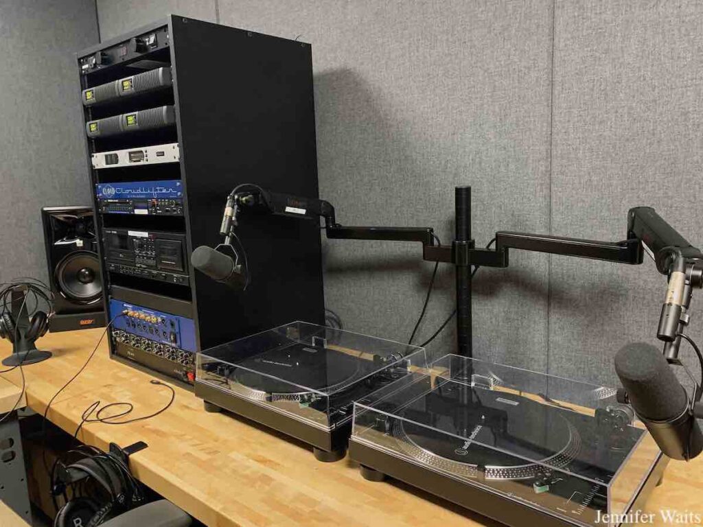 Photo of studio at college radio station WONY-FM. Pictured are two turntables with two microphones above them. To the left is a rack of audio equipment, with a speaker to the left of that. Headphones are attached to a stand and to a hook on the furniture. Photo: J. Waits