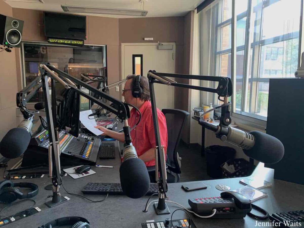 Photo of male DJ wearing headphones and holding a piece of paper while talking on a microphone during his radio show. In the college radio studio are multiple microphones, a mixing board, phone, headphones, speakers, and a computer monitor. Photo: J. Waits