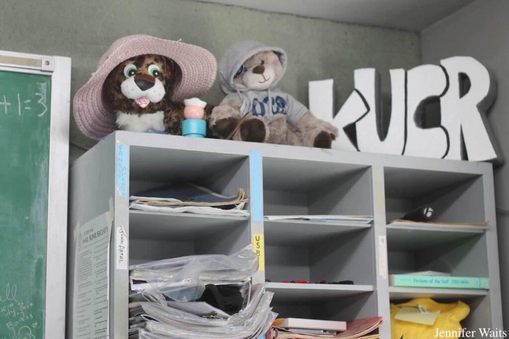 Photograph of grey shelves that serve as mailboxes for college radio station KUCR. On top of the shelves are two stuffed animal bears and a sign that depicts the letters K-U-C-R. Photo: J. Waits