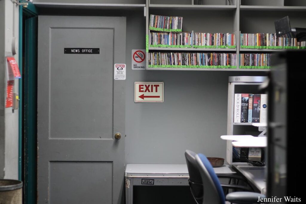 Photo of room at college radio station KUCR. "News Office" sign is on a grey door that is open. CDs are on upper shelves and a black file cabinet is in the foreground, next to desks with office chairs. Photo: J. Waits