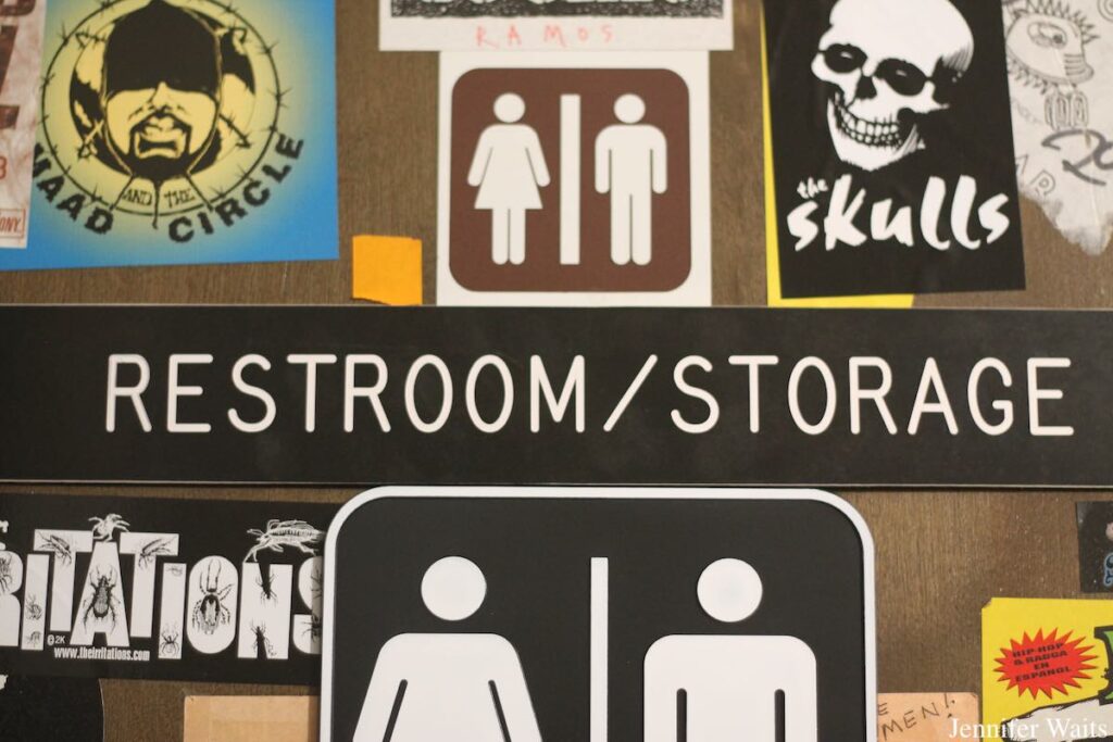 Photo of sticker-covered restroom door at UC Riverside's college radio station KUCR. In the center of the photo is a sign that reads "restroom/storage." Above and below those words are images of male and female icons. Bands stickers are affixed all over the door, including a sticker for The Skulls. Photo by J. Waits