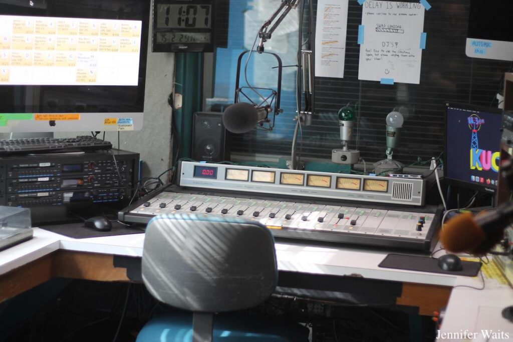 Photograph of studio at UC Riverside's college radio station KUCR. An office chair is in the foreground, with a mixing board in the center. Audio equipment and computer monitors are also pictured, as well as a clock that reads 11:07. Photo: J. Waits