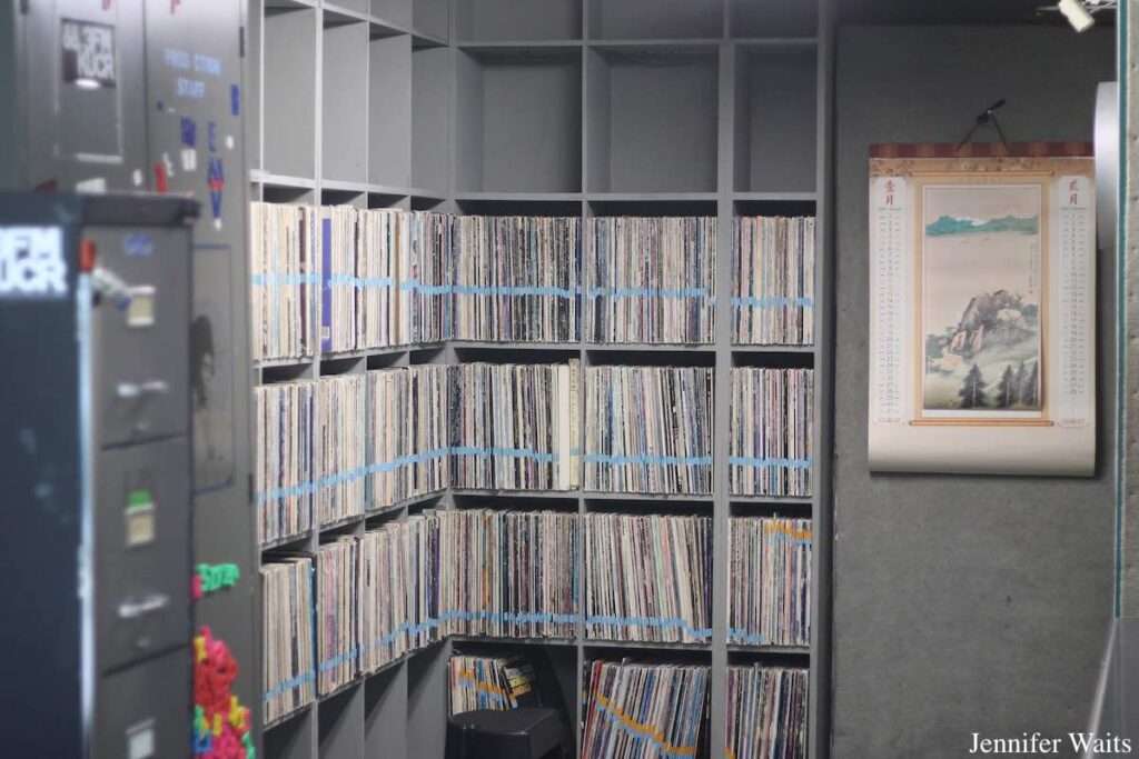 Photograph of room at college radio station KUCR. The walls are grey and shelves hold 12" vinyl records. A calendar is on the wall to the rights and a file cabinet is to the left of the vinyl. Photo: J. Waits