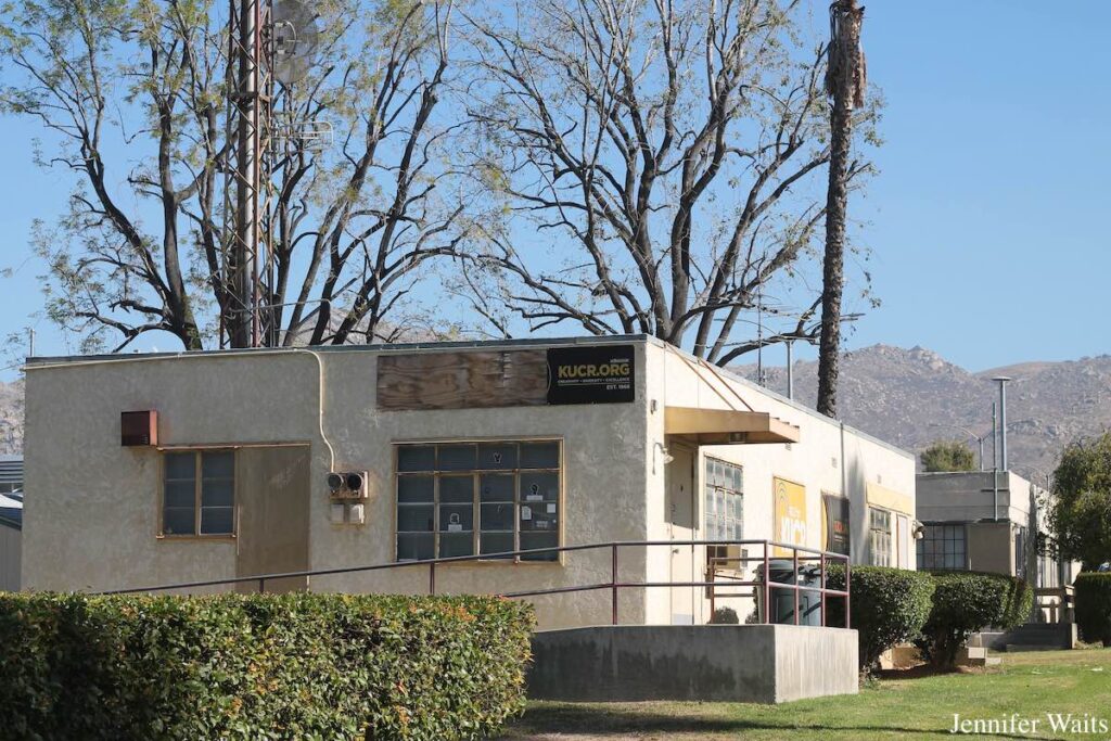 Photo of boxy white stucco building, with wheelchair ramp and railing on one side leading to an entrance door. KUCR.org is written on a sign on the building. Trees can be seen behind the building, with mountains in the distance. Photo: J. Waits